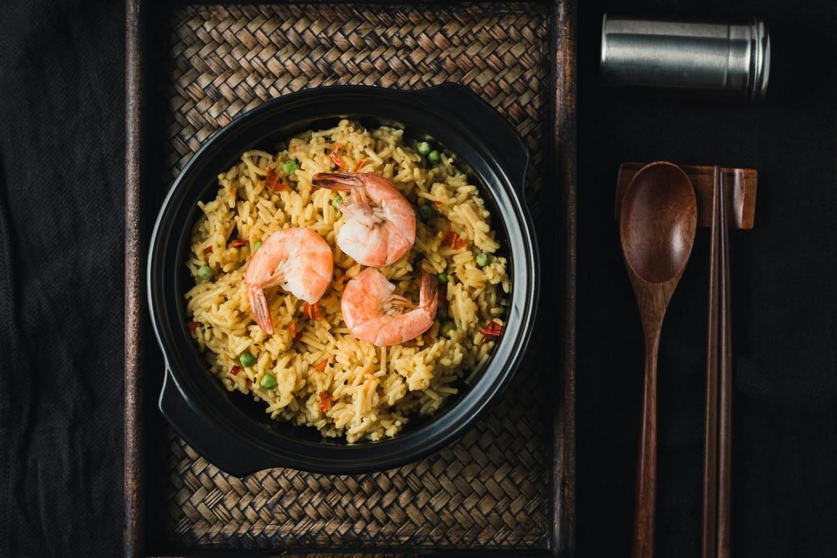 A top-down view of a black ceramic bowl filled with seasoned yellow rice, green peas, and three large cooked shrimp. The dish rests inside a woven bamboo tray alongside wooden chopsticks, a wooden spoon, and a metal shaker on a dark background.