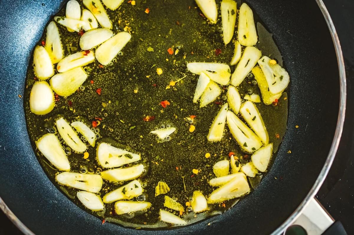 Overhead close-up shot of sliced garlic simmering in oil with chili flakes inside a black frying pan, highlighting texture, aroma, and the start of a flavorful cooking process.