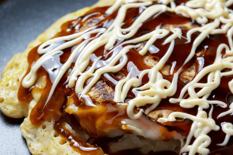 A close-up of crispy okonomiyaki drizzled with thick brown sauce and creamy mayonnaise in a decorative swirl—Japan’s beloved savory pancake, hot off the griddle and ready to savor.