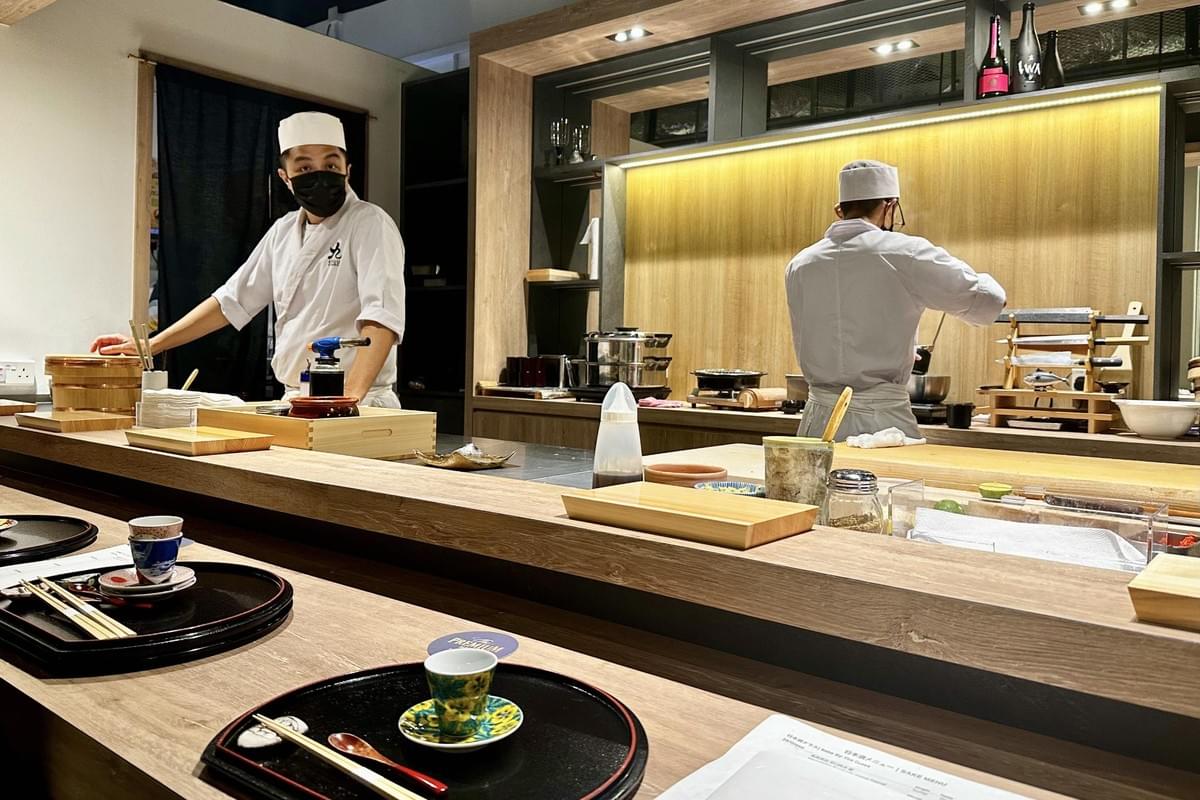 This image captures a modern sushi counter where two chefs in traditional white uniforms and caps are at work behind a wooden bar. The scene is meticulously arranged with individual place settings, lacquer trays, and various culinary tools under warm, recessed lighting.