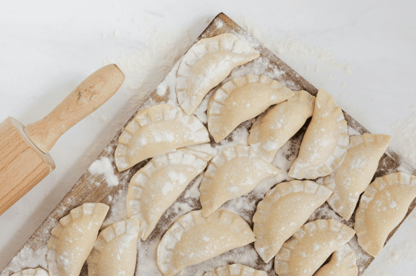 A batch of freshly handmade, uncooked pierogi with classic crimped edges arranged on a floured wooden board next to a rolling pin.