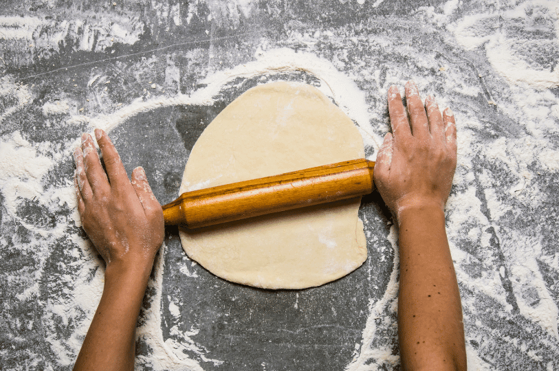 Overhead shot of hands using a wooden rolling pin to flatten pierogi dough on a flour-dusted grey countertop.