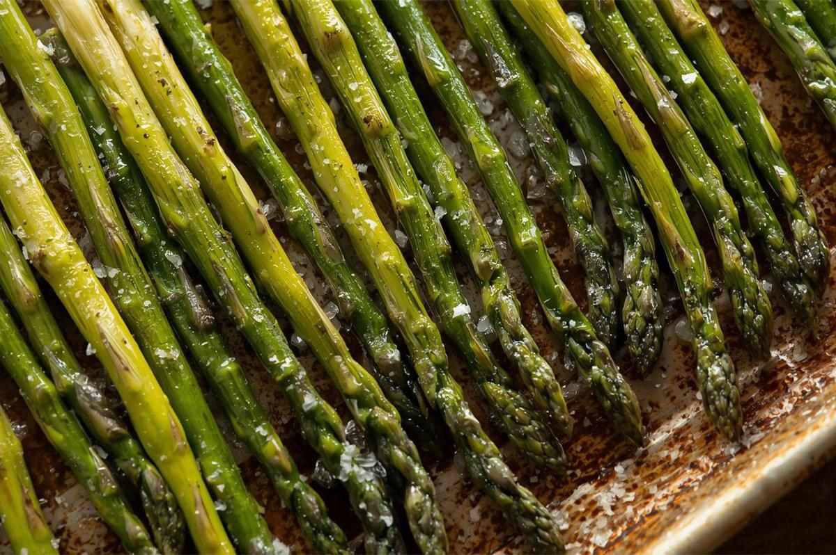 A row of bright green asparagus spears evenly lined up on a baking sheet, glistening with olive oil and sprinkled generously with coarse sea salt before or after roasting.