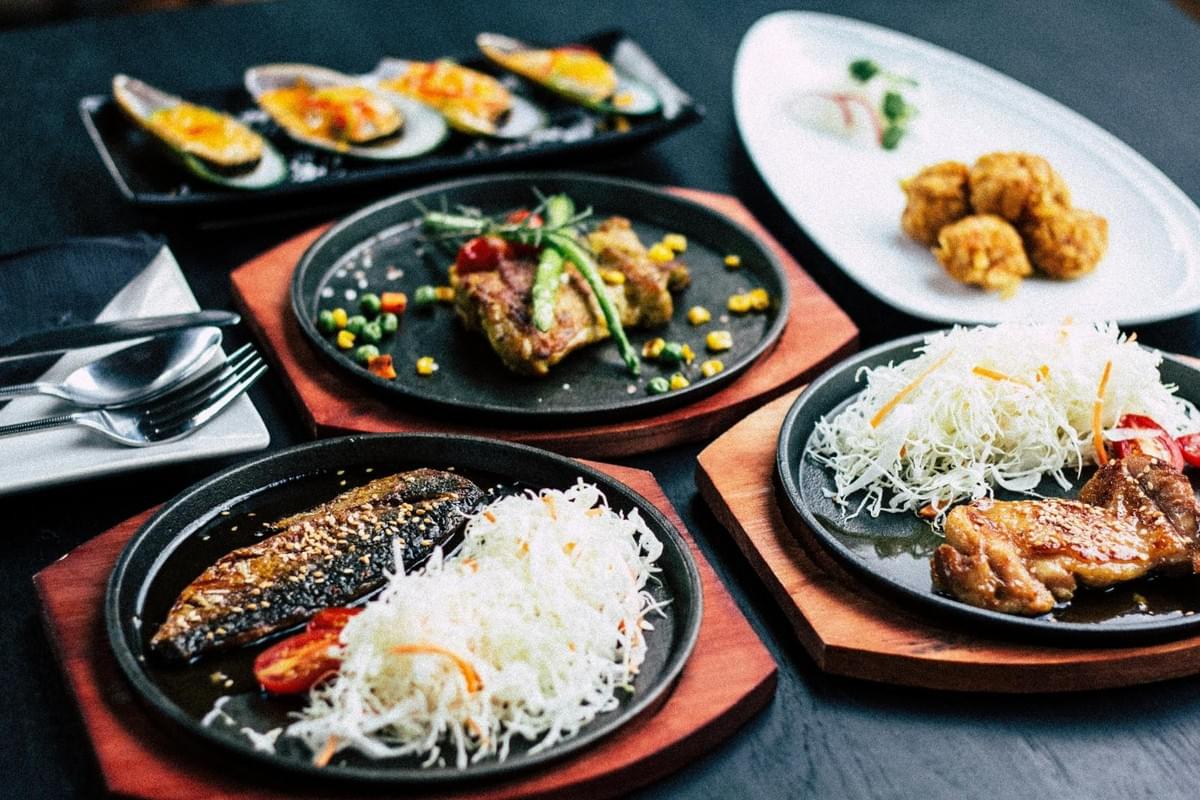 A tabletop array of Asian-style dishes features sizzling hot plates of glazed grilled chicken, grilled mackerel, and chicken breast garnish, each accompanied by shredded cabbage and vegetables. Surrounding the hot plates are side dishes, including baked cheese-topped mussels in half shells and crispy fried meatballs served on white porcelain plates. Midjour