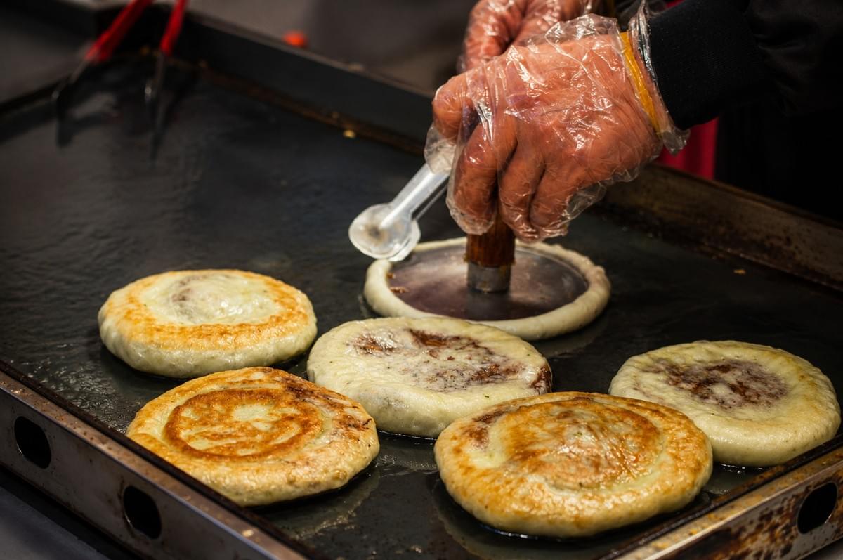 A vendor pressing dough with a flat tool while cooking hotteok pancakes on a large hot griddle.