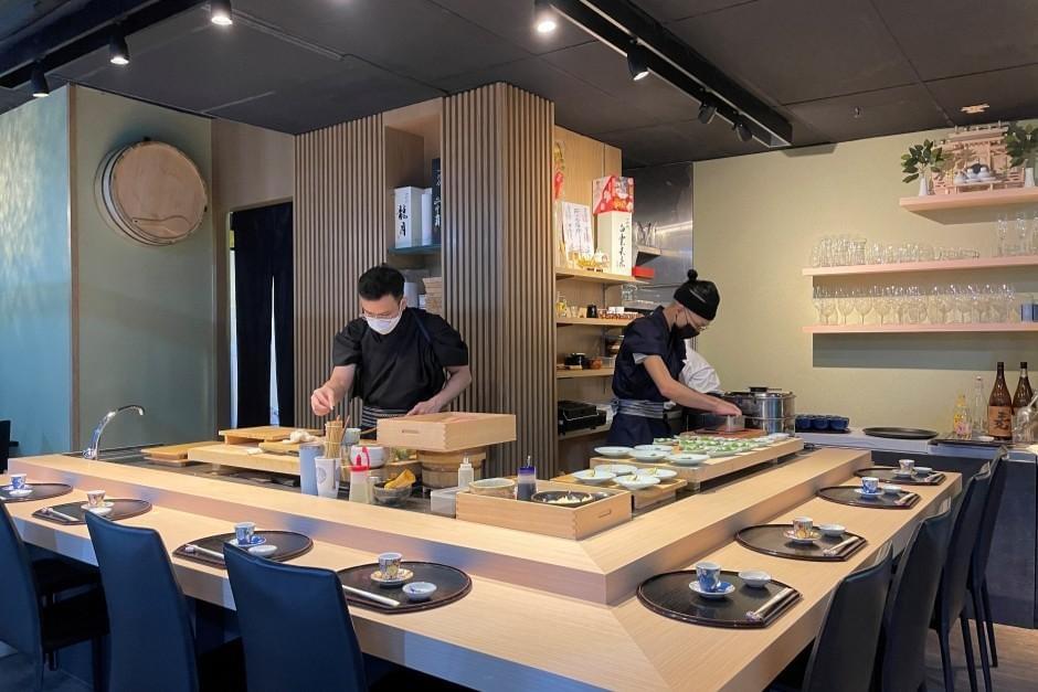 This image captures a modern sushi counter where two chefs in traditional white uniforms and caps are at work behind a wooden bar. The scene is meticulously arranged with individual place settings, lacquer trays, and various culinary tools under warm, recessed lighting.
