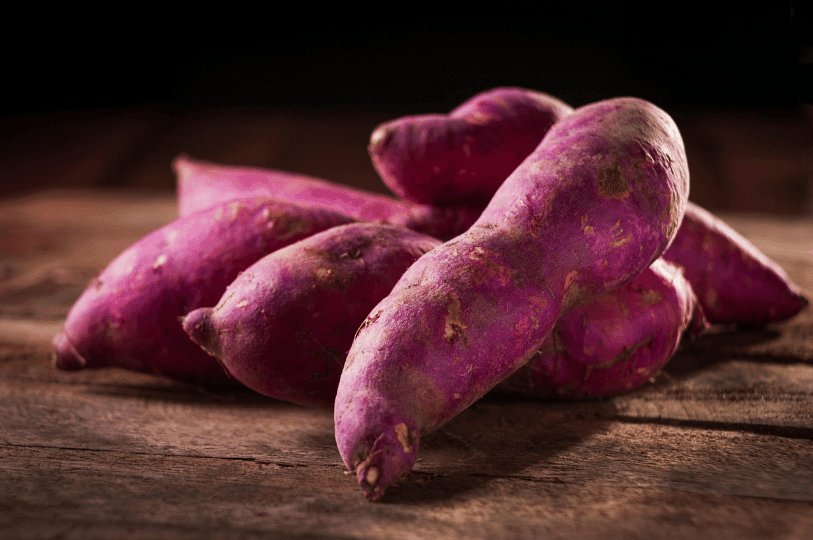 A group of raw, purple-skinned sweet potatoes piled on a rustic wooden surface against a dark background, highlighting their natural texture and earthy appearance.
