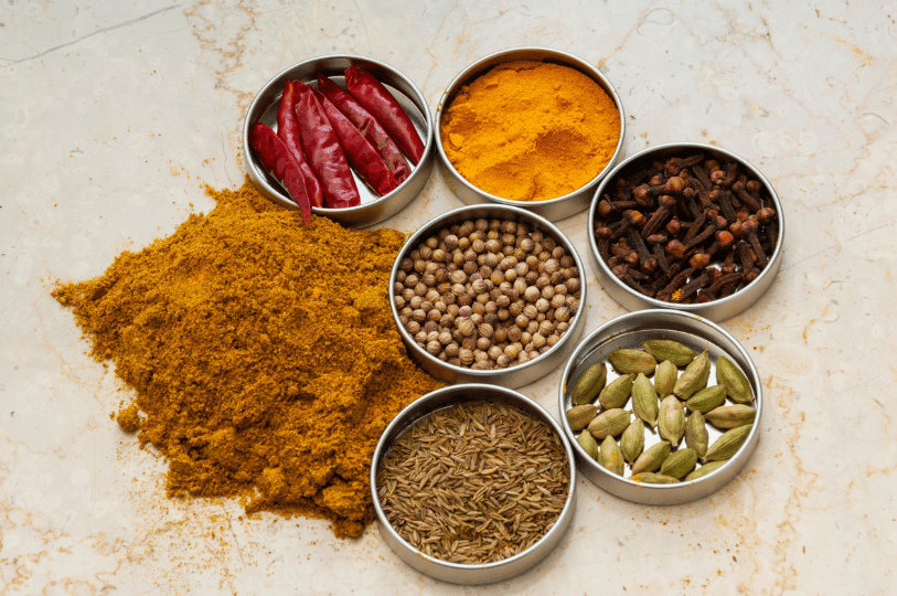 A flat-lay view of a culinary workspace featuring a large pile of vibrant yellow curry powder next to small metal tins containing whole dried red chilies, turmeric powder, cloves, green cardamom pods, cumin seeds, and coriander seeds on a marble surface.