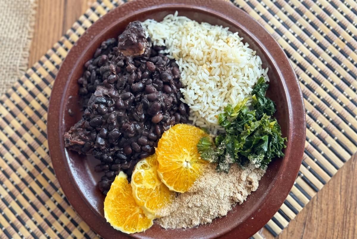 Earthenware plate filled with black beans, white rice, sauteed greens, toasted cassava flour, and orange slices.