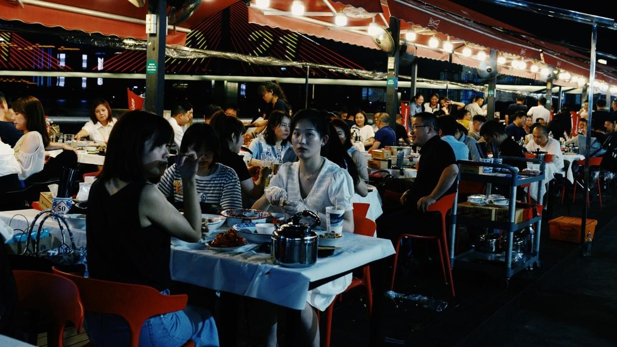 Under the warm glow of string lights and red canopies, a bustling outdoor restaurant is packed with diners enjoying a meal at white-clothed tables. The scene is set against a night backdrop featuring an illuminated suspension bridge, with a woman in the center foreground looking directly into the camera lens.