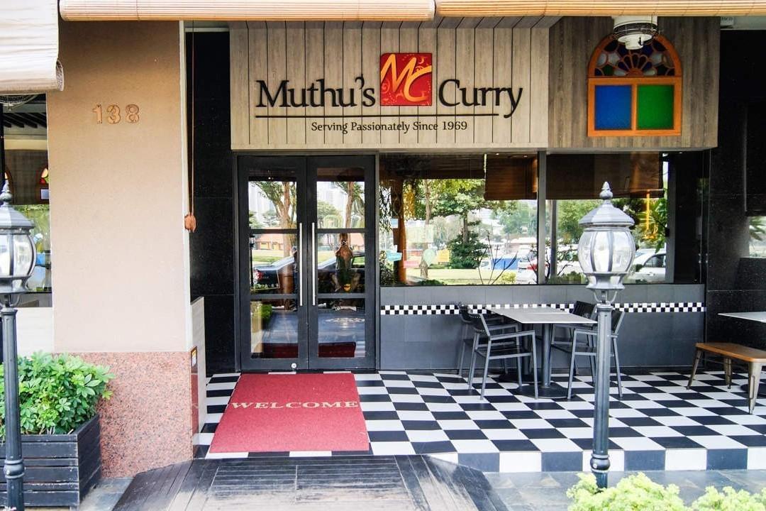 The image shows the storefront of a restaurant named "Muthu's Curry," featuring double glass entrance doors, a red welcome mat, and outdoor seating on a black-and-white checkered floor. The prominent signage above the door includes the restaurant's name and the tagline, "Serving Passionately Since 1969."