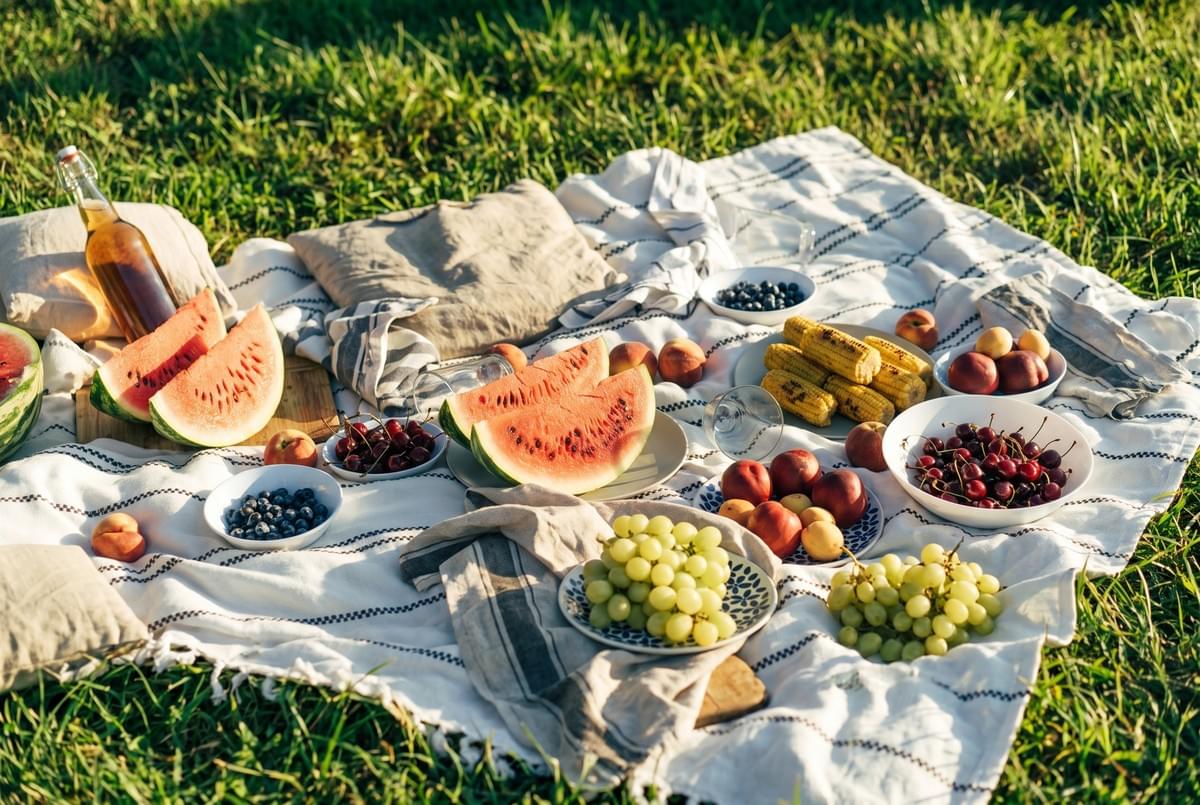 Flat lay of a picnic blanket with fresh watermelon slices, grapes, peaches, blueberries, and grilled corn cob halves.