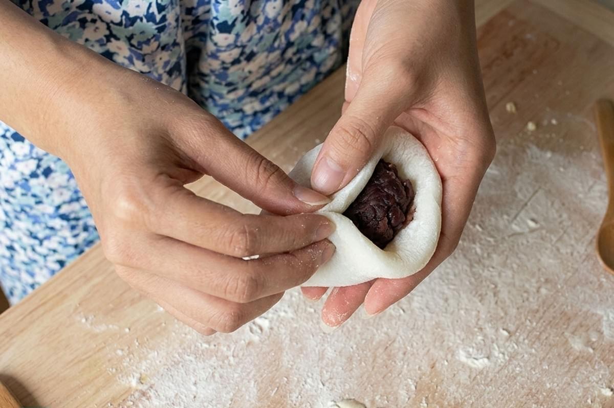 Hands wrapping mochi dough around red bean paste filling on wooden board