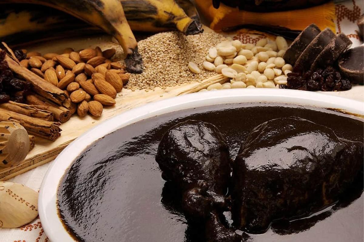 Dramatic still life of a plate of chicken smothered in rich, dark Mole Negro, surrounded by the ingredients used to make the sauce, including chocolate, almonds, spices, and plantains.