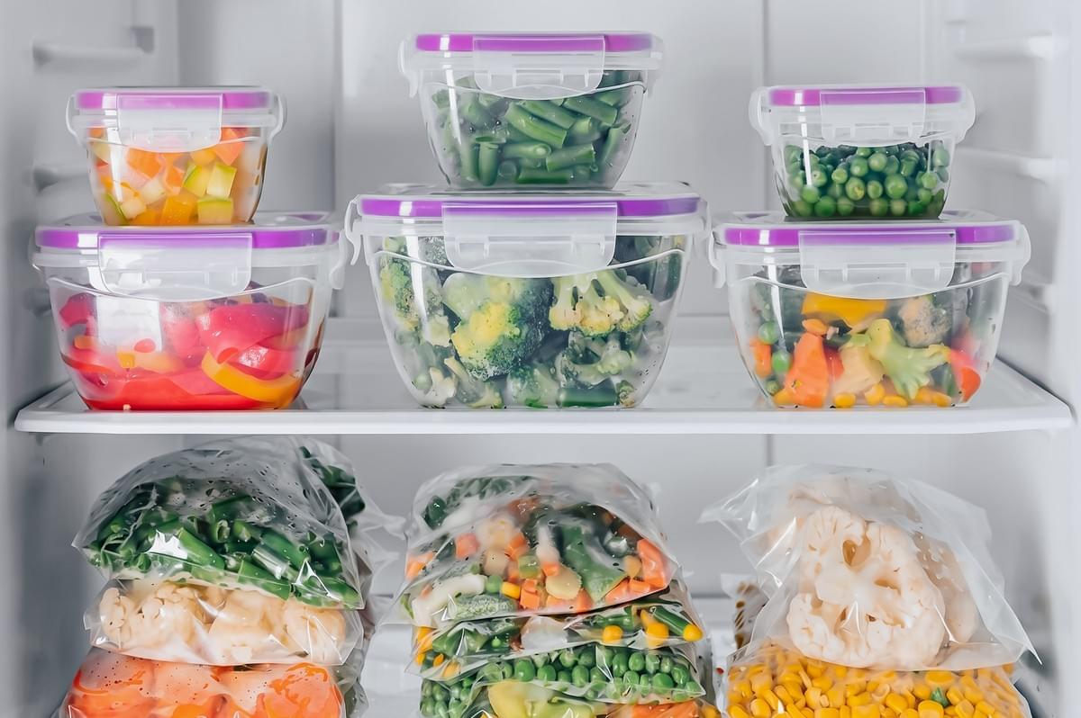 Straight-on wide shot inside a refrigerator showing clear food storage containers and freezer bags filled with mixed vegetables, highlighting organized fridge storage and freezing techniques to reduce food waste and maintain food safety.