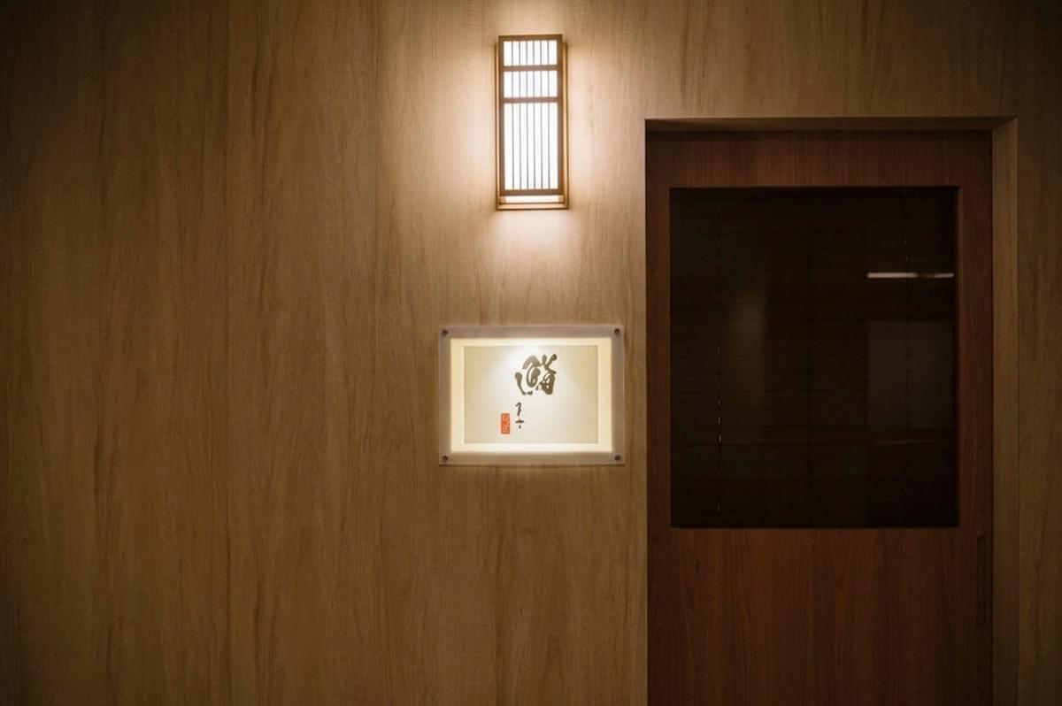 A minimalist shot of a restaurant entrance featuring a light-toned vertical wood-paneled wall. A simple, rectangular wooden door with a dark glass pane is on the right. To the left of the door, a backlit rectangular sign displays artistic Japanese calligraphy. Above the sign, a traditional-style rectangular lantern with vertical wooden slats casts a warm, inviting glow downward.