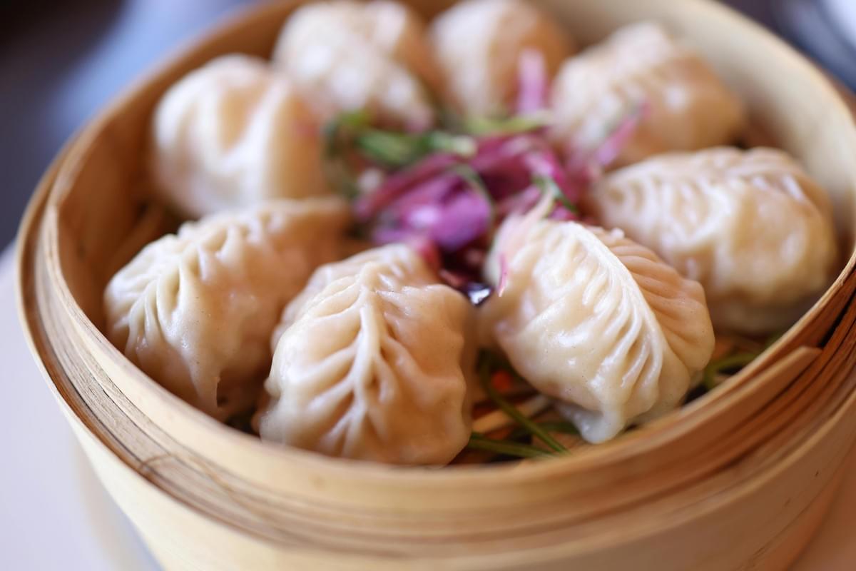 A close-up shot shows seven steamed dumplings arranged in a circular bamboo steamer basket. The plump, pleated dumplings surround a central garnish of bright purple microgreens.