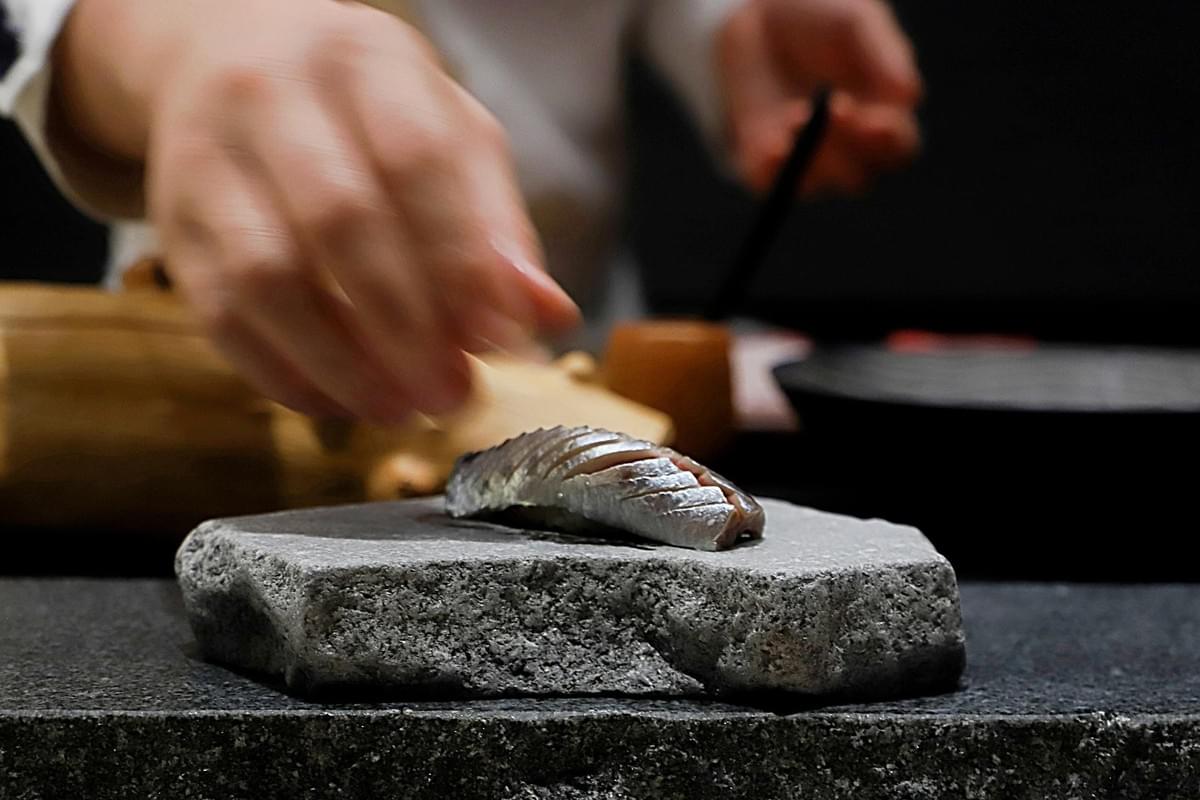 A single piece of expertly sliced nigiri sushi rests on a rustic, grey stone slab, highlighting its delicate texture. In the blurred background, a chef’s hands move with precision to finish the presentation of the dish.