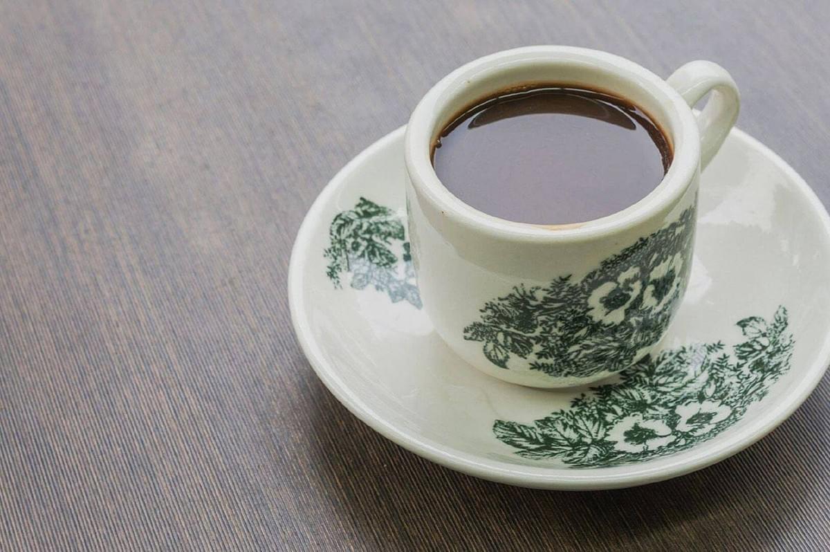 A white cup filled with dark coffee sits on a matching saucer, both adorned with green floral designs, placed on a light wood surface. The tone is cozy and inviting.