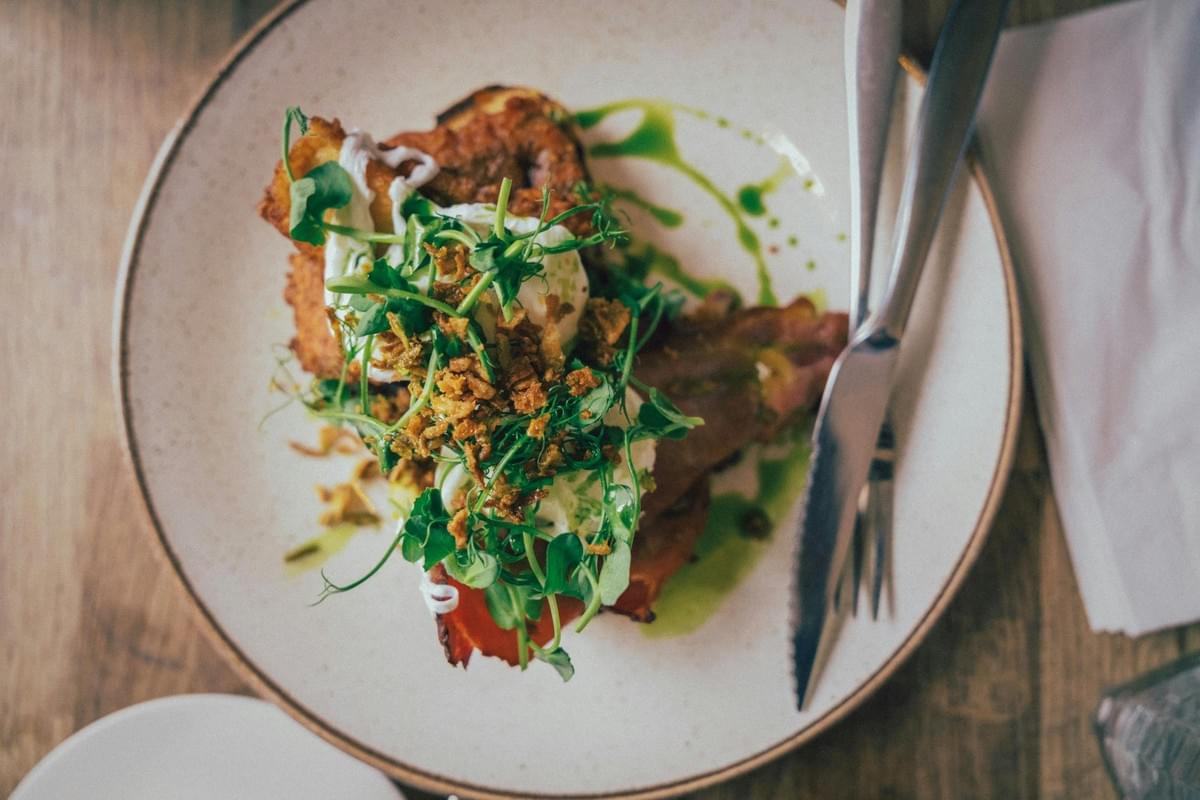 A plated gourmet breakfast dish featuring toast, bacon, and poached eggs rests on a round ceramic plate next to silver cutlery. The meal is topped with fresh microgreens, crispy fried onions, and a drizzle of green herb oil.
