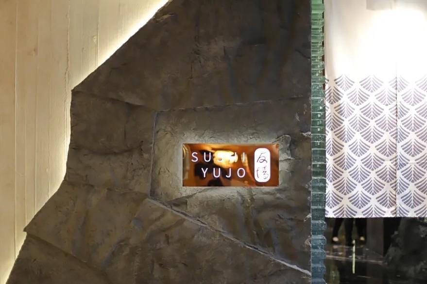 An illuminated sign reading "SUSHI YUJO" is embedded within a textured gray rock facade with backlighting along its upper edge. To the right hanging adjacent to the stone feature is a white curtain patterned with subtle blue leaf motifs.