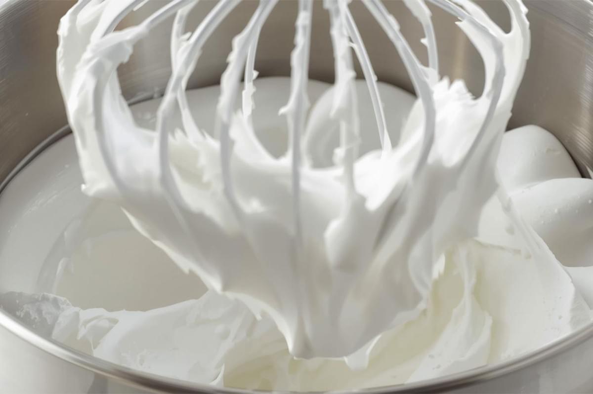 Close-up of a wire whisk attachment in a stainless steel stand mixer bowl, whipping a thick, glossy white mixture (likely meringue or marshmallow base) to stiff peaks.