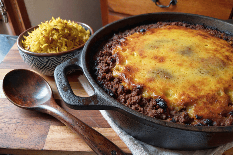 A hearty portion of bobotie in a black cast-iron skillet featuring a bubbly, browned custard topping, served with a bowl of fluffy yellow rice.