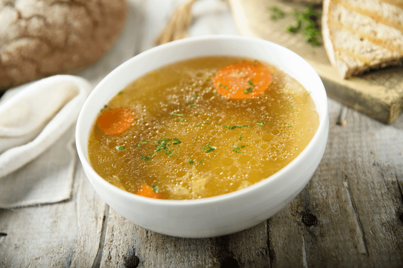 A white bowl of clear, golden chicken broth containing sliced carrots and fresh herbs, served on a rustic wooden table with bread in the background.