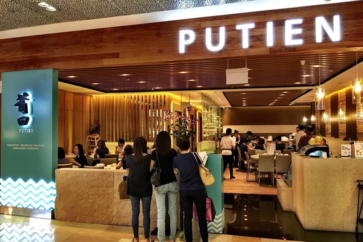 The image shows the bright storefront of a Putien restaurant, featuring a large illuminated sign and warm wood paneling. Inside, several patrons are seated at tables or waiting near the entrance of the modern, well-lit dining area.