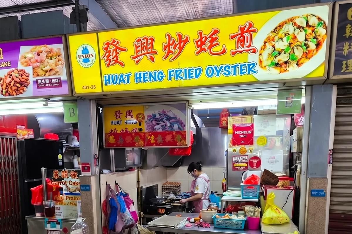 The storefront of Huat Heng Fried Oyster in a Singapore hawker center. A vendor in a pink apron works at the griddle inside the small stall, which is decorated with Michelin Guide recognition stickers and traditional signage.