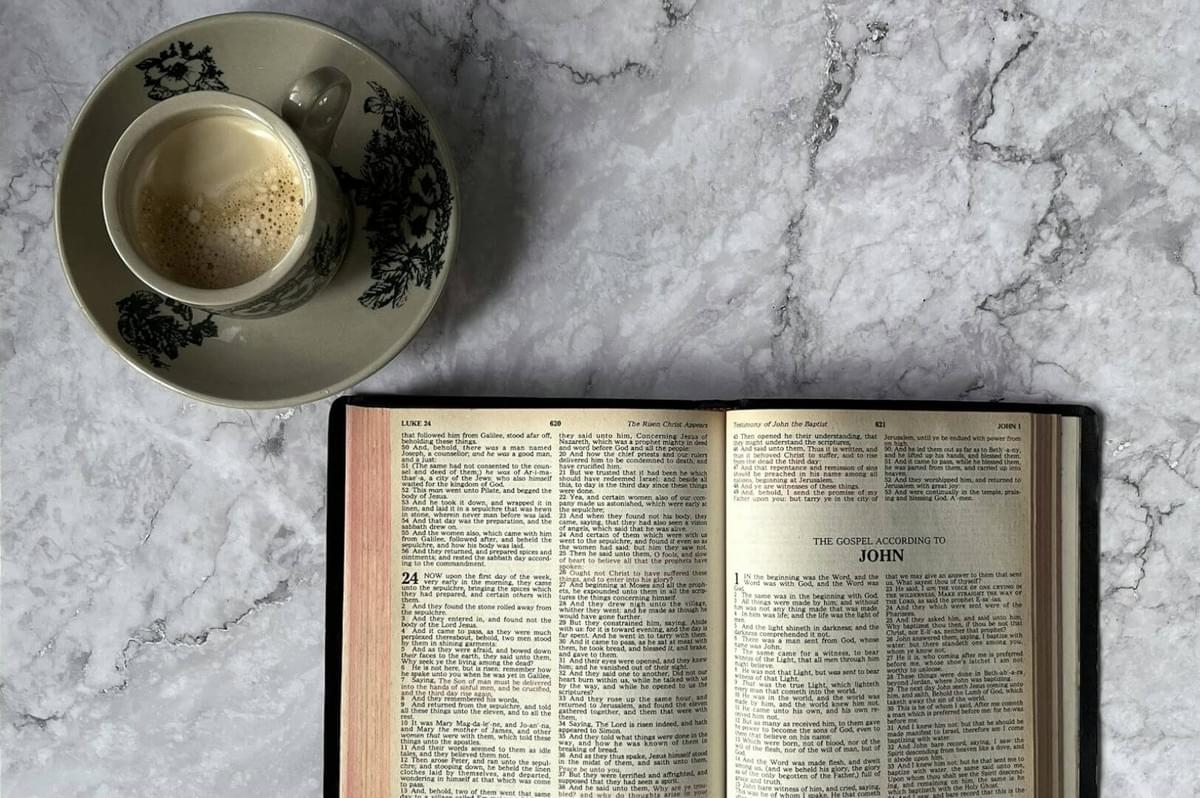 A cup of frothy coffee sits on a floral saucer beside an open Bible on a marble surface. The scene is calm and inviting, suggesting quiet reflection.