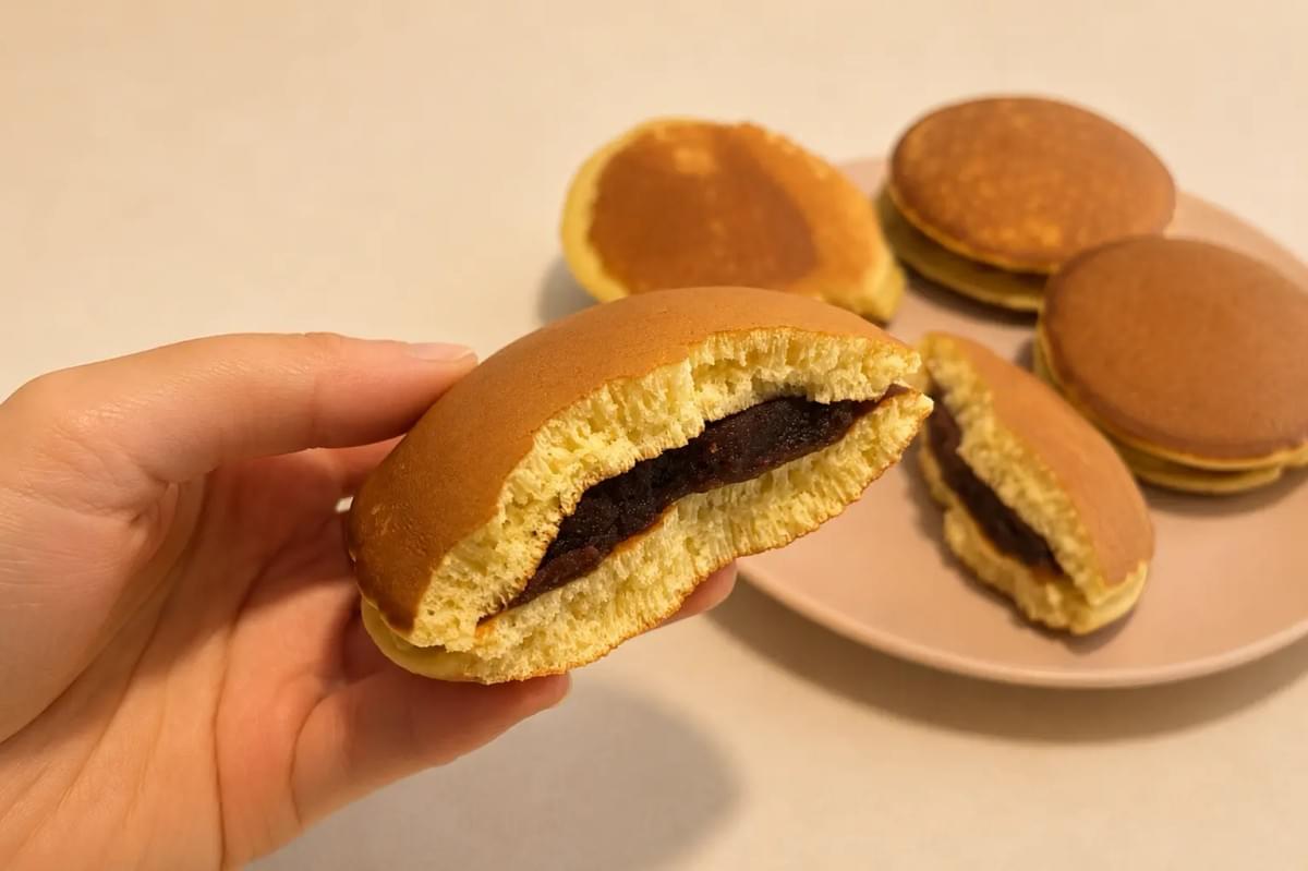 A close-up shot of a hand holding a dorayaki pancake sandwich with a large bite taken out, showcasing the thick sweet red bean paste inside the fluffy, golden interior. In the background, more whole and halved dorayaki sit on a light pink plate against a neutral tabletop.