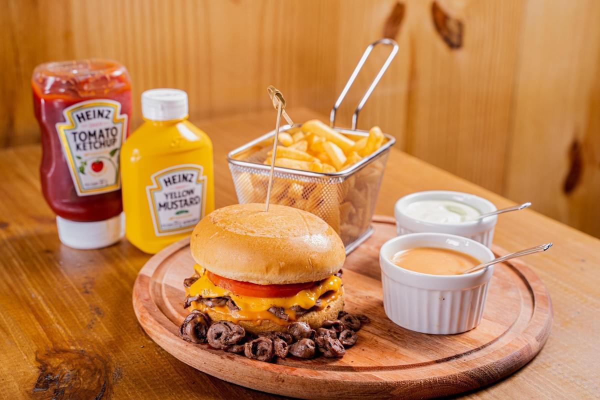A cheeseburger with a tomato slice and unique curly meat toppings is served on a round wooden board alongside a wire basket of french fries and two small dipping cups. In the background on a wooden table, plastic squeeze bottles of Heinz tomato ketchup and yellow mustard are visible.