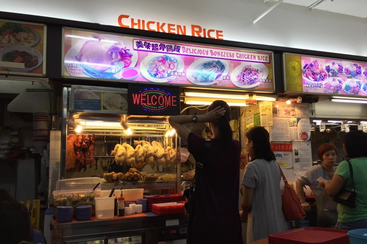  The image displays a bustling food stall featuring a "Delicious Boneless Chicken Rice" sign with photos of various dishes like steamed chicken and oyster sauce vegetables. Customers stand at the counter where whole cooked chickens hang in a glass display case under a bright "WELCOME" sign.
