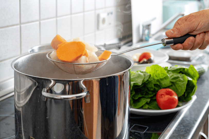 Sliced carrots and root vegetables being removed from boiling water using a mesh strainer, ready to be transferred to an ice bath.