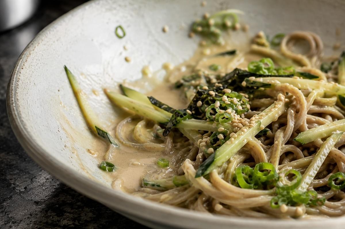 A tight, detailed close-up focuses on a portion of the cold soba noodles after they have been mixed with the creamy sesame-tahini sauce in a shallow ceramic bowl. The glossy, tan sauce coats the individual strands of buckwheat noodles, interspersed with vibrant green julienned cucumbers and chopped scallions. Toasted sesame seeds and bits of dark nori are scattered throughout the mixture, with remnants of the rich sauce smeared along the textured inner rim of the bowl.