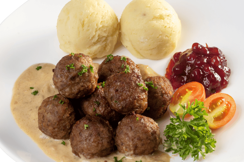A close-up of several meatballs drizzled with creamy brown gravy, served with two neat scoops of mashed potatoes, lingonberry sauce, sliced cherry tomatoes, and a sprig of curly parsley on a bright white plate.