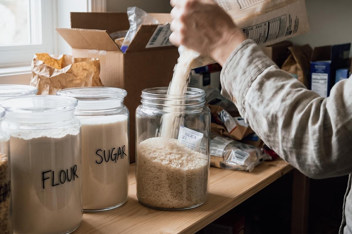 Close-up side angle shot of dry rice being poured into a glass airtight container with a silica gel packet, illustrating pantry storage solutions to prevent moisture and extend shelf life in humid climates.