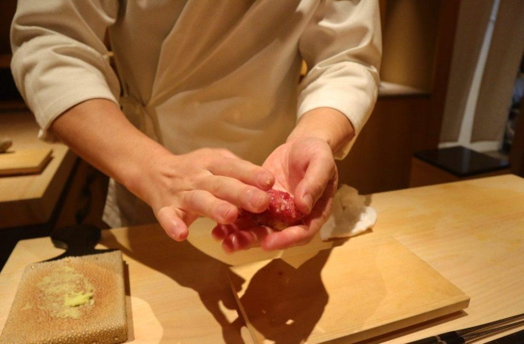 A sushi chef wearing a white chef's jacket carefully shapes a piece of nigiri with both hands over a wooden counter. To the left, a traditional sharkskin grater holds a small mound of freshly grated wasabi.
