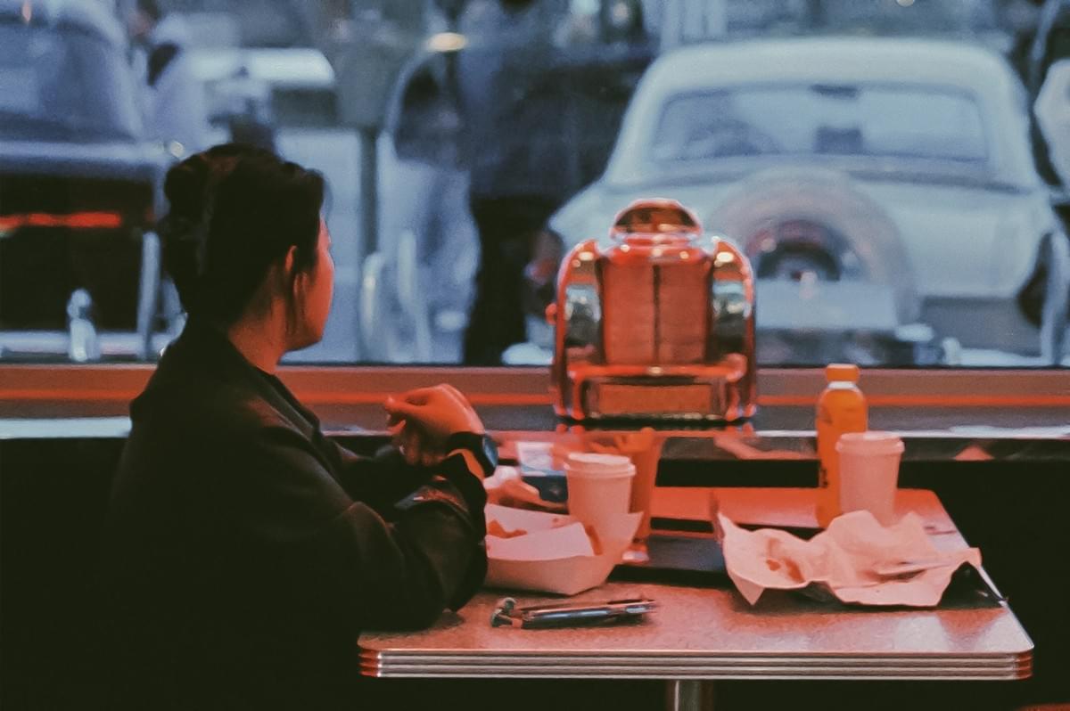 Wide-angle eye-level shot of a person seated at a retro-style diner counter with takeaway food and drinks on a metal table, looking out through a large window at parked cars, creating a nostalgic urban dining atmosphere.