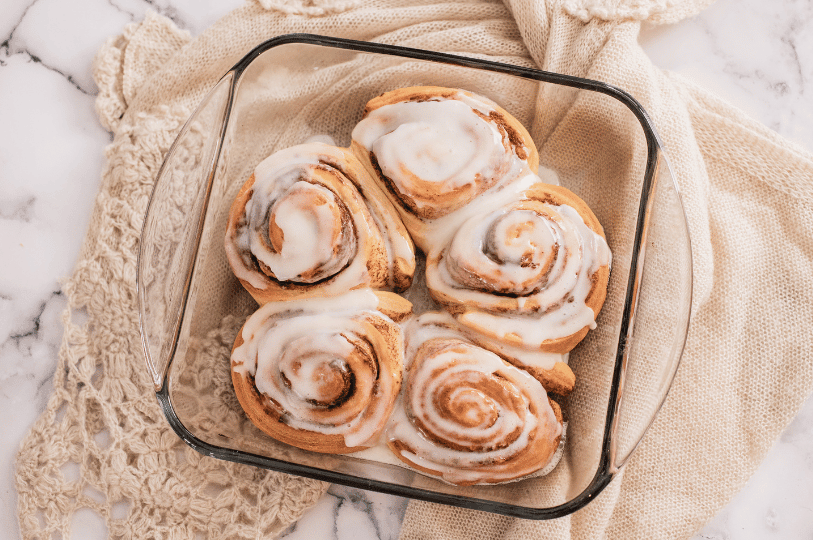Top-down view of five large, fluffy cinnamon rolls nestled in a square glass baking dish, generously topped with a smooth sugar glaze.