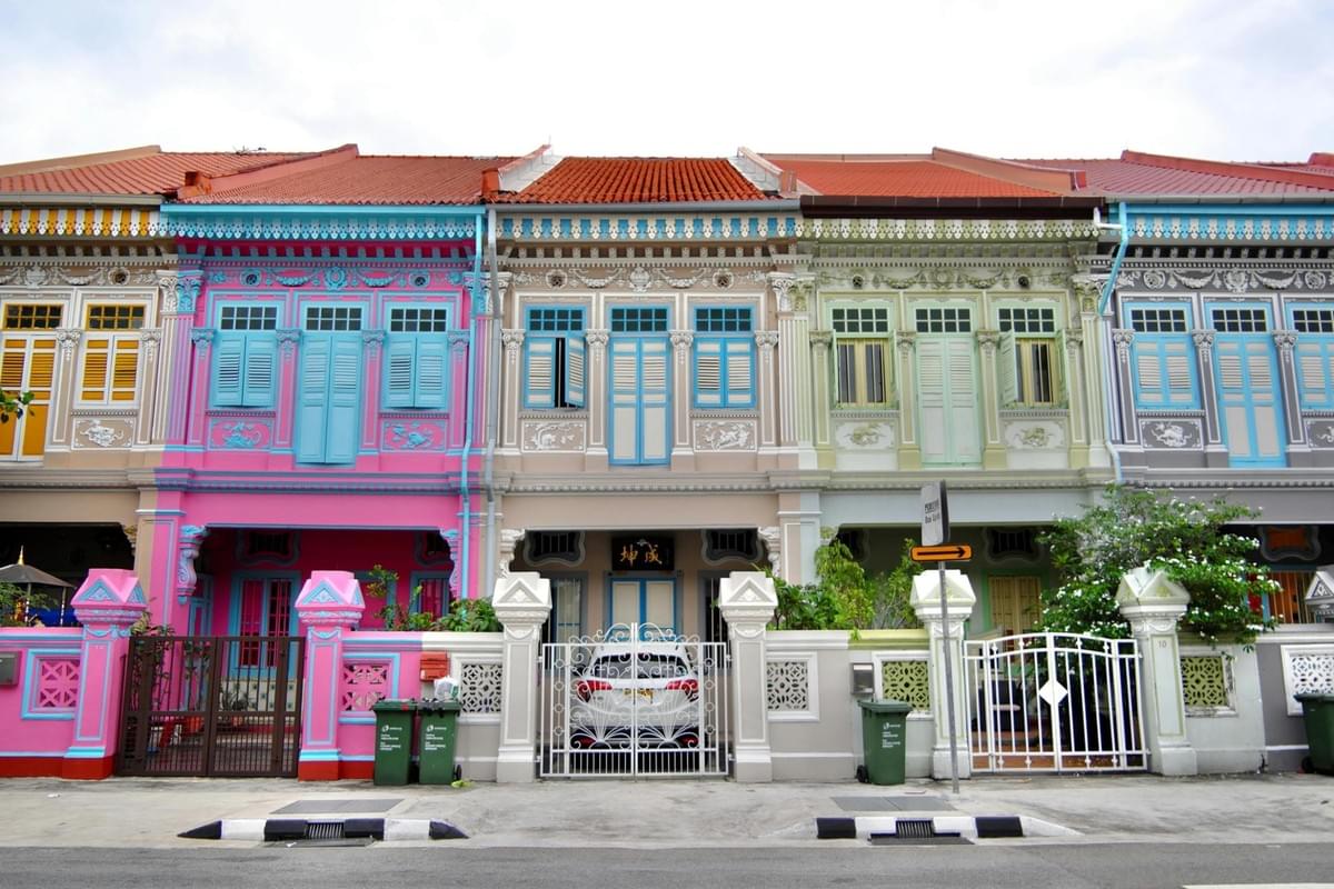 This image showcases a row of vibrant, multi-colored Peranakan-style terrace houses featuring intricate architectural details and traditional window shutters. Each house is painted in a distinct pastel or bold hue—ranging from bright pink to soft mint—and is fronted by decorative gates and low fences.
