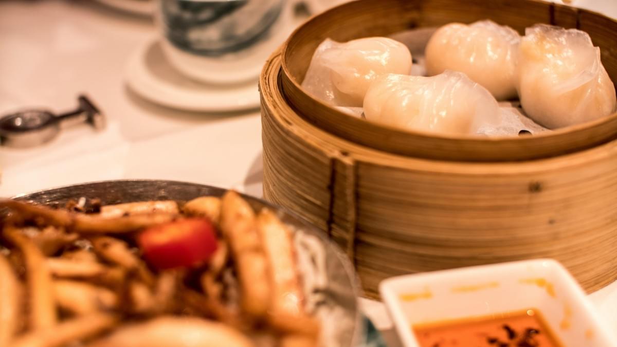 This is a close-up, shallow depth-of-field shot of food on a white tablecloth, prominently featuring a bamboo steamer basket filled with translucent white dumplings, likely Har Gow. In the blurry foreground.