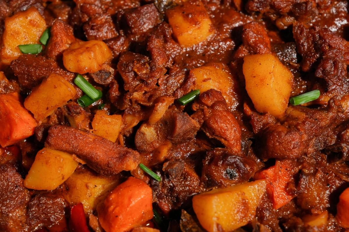 This close-up captures a hearty dish of pork afritada, featuring tender chunks of meat, potatoes, and carrots coated in a rich, reddish-brown sauce. Fresh green onion garnishes are scattered across the top, providing a vibrant color contrast to the savory, glossy texture of the stew.