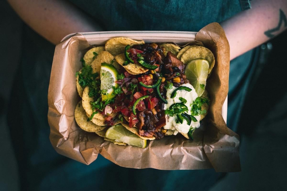 A person holds a paper tray filled with loaded nachos topped with black beans, salsa, jalapeños, and a dollop of cream. Fresh lime wedges and chopped cilantro garnish the dish, which is served over brown parchment paper.