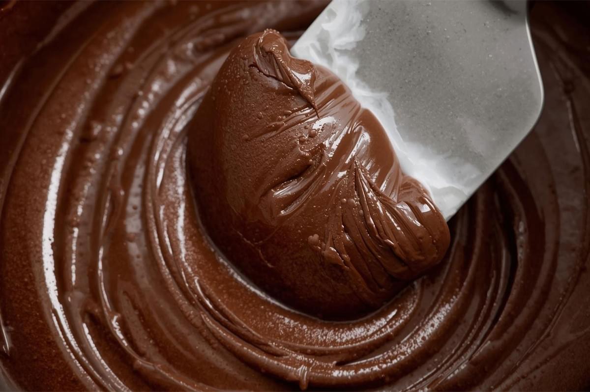 Extreme close-up of thick, smooth, melted chocolate or ganache being swirled with a white spatula in a bowl.