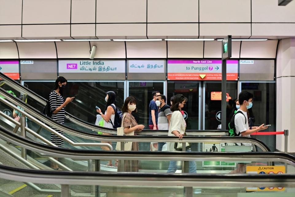 This image captures commuters traveling on escalators within the Little India MRT station, clearly identified by the overhead purple signage for the North East Line towards Punggol.