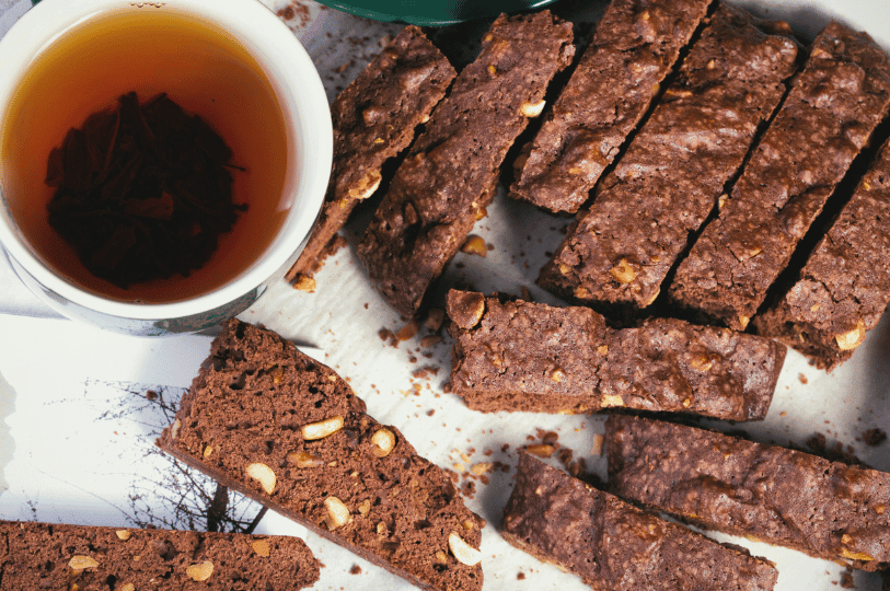 Chocolate biscotti with nutty chunks rest beside a steeping cup of tea, their rich texture and deep brown hue inviting a cozy, twice-baked indulgence.