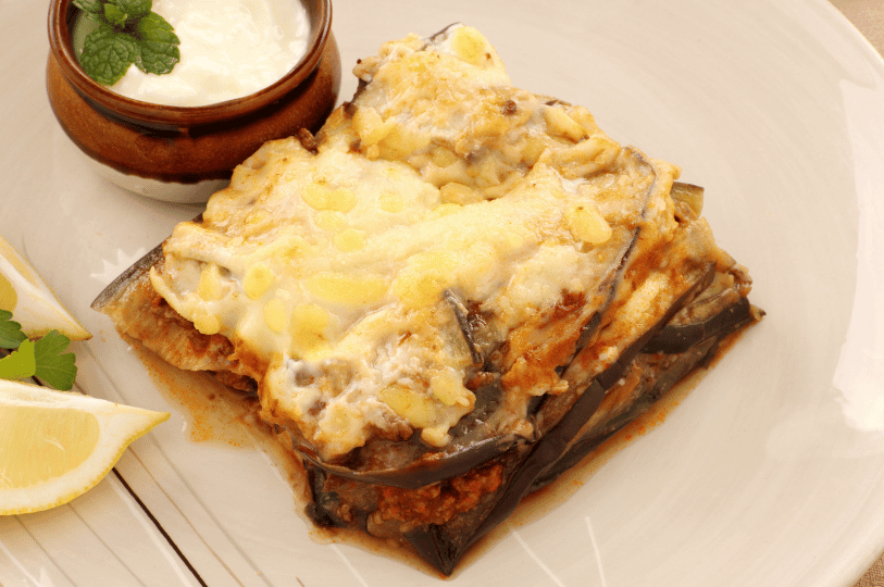 A golden-baked serving of Greek moussaka with creamy béchamel and layered eggplant and meat, plated with lemon wedges, parsley, and mint-garnished yogurt—offering a rich, comforting taste of the Mediterranean.
