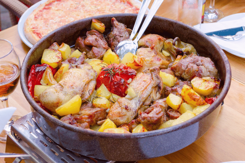 A round pan of traditional Croatian meat Peka featuring golden roasted chicken pieces, potatoes, green bell peppers, and a whole roasted tomato in the center, resting on an outdoor table.